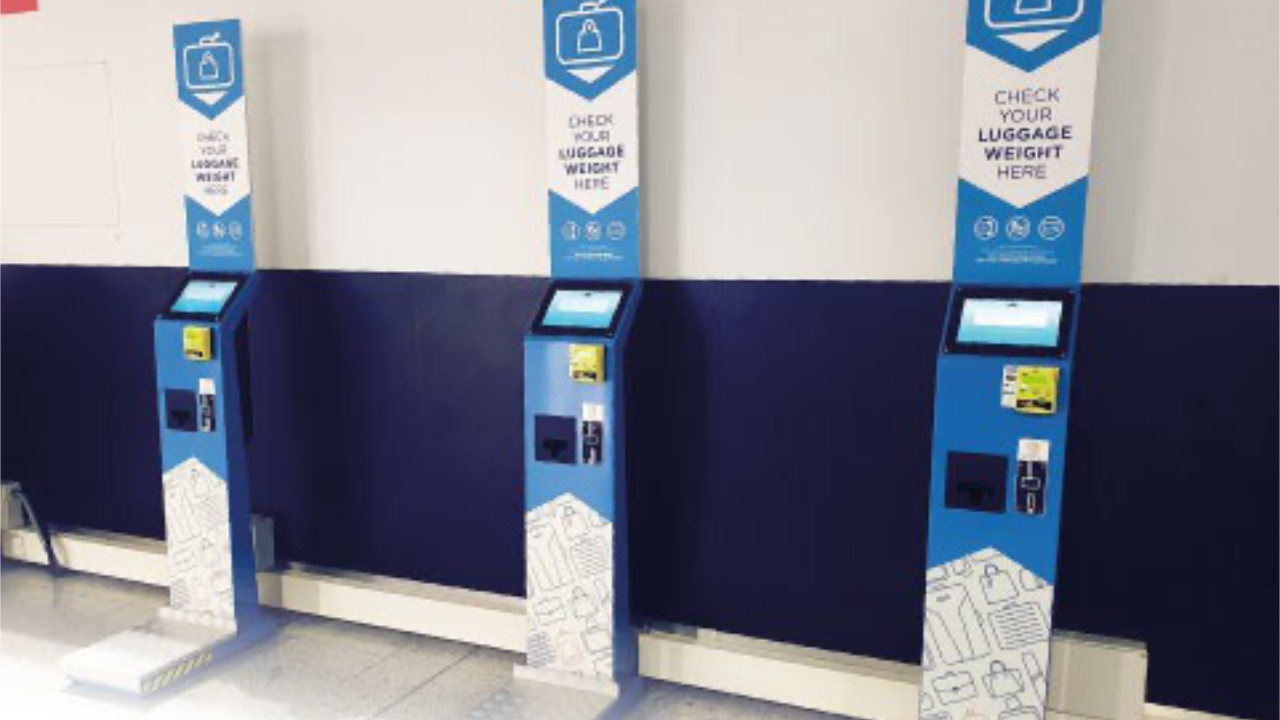Self-service kiosks for checking luggage weight lined up against a wall in an airport or travel facility